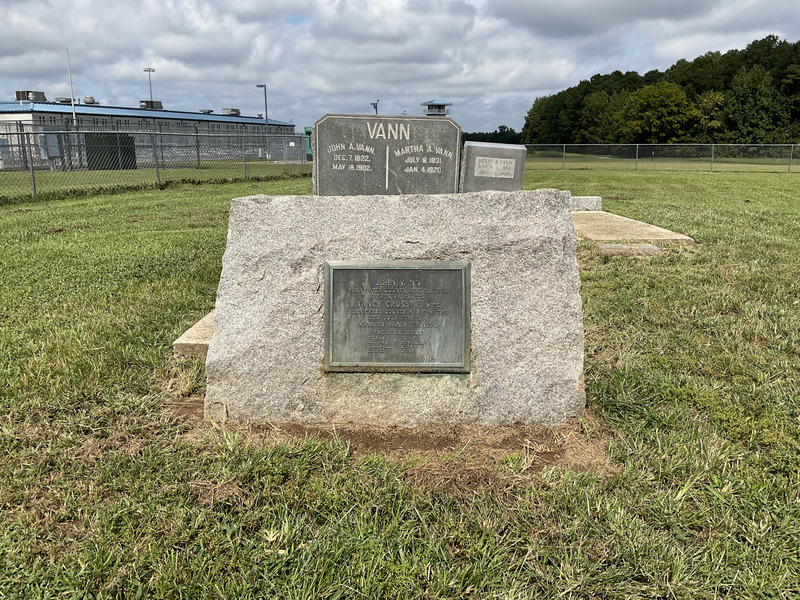 Tombstones of John Vann and his wife, Nancy Cross, with the prison watchtower to the rear. The prison watchtower casts a shadow on the landscape of the nine-tombstone plantation-era cemetery connected to the main road by a narrow dirt and gravel road. 