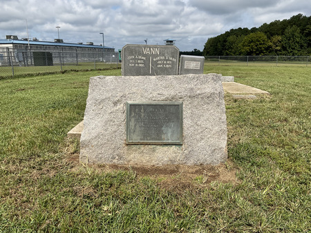Tombstones of John Vann and his wife, Nancy Cross, with the prison watchtower to the rear. The prison watchtower casts a shadow on the landscape of the nine-tombstone plantation-era cemetery connected to the main road by a narrow dirt and gravel road. 