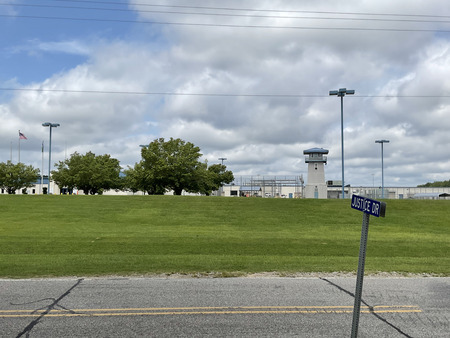 The blue street sign with "Justice Dr" sits across from the gated RCI facility and watchtower.